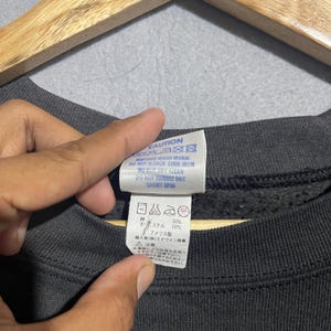 Op de afbeelding: Close-up van een donkergrijs sweatshirtlabel met wasvoorschriften. Het label leest "CAUTION" en bevat wassymbolen. Het label vermeldt ook de samenstelling van de stof als 50% polyester en 50% katoen. Het label wordt door een hand vastgehouden.