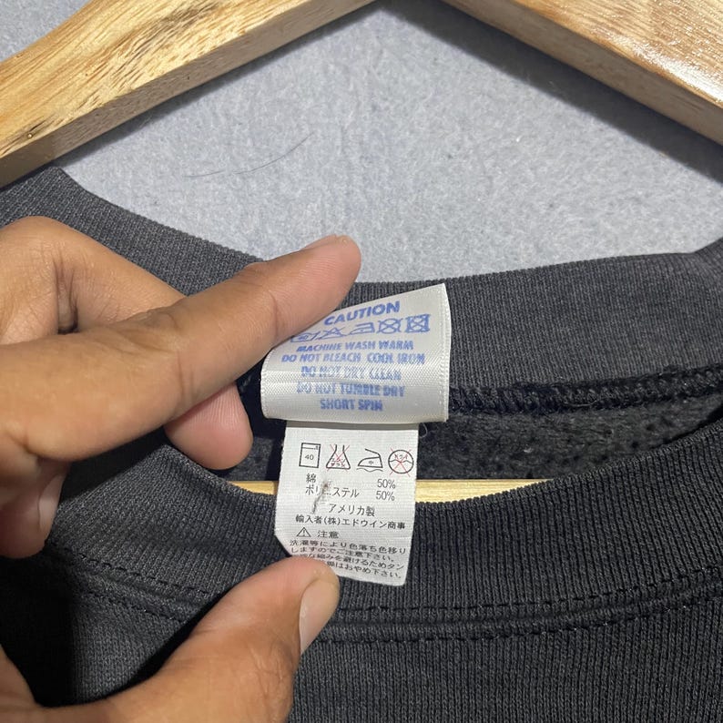 Op de afbeelding: Close-up van een donkergrijs sweatshirtlabel met wasvoorschriften. Het label leest "CAUTION" en bevat wassymbolen. Het label vermeldt ook de samenstelling van de stof als 50% polyester en 50% katoen. Het label wordt door een hand vastgehouden.