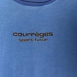 Puede incluir: Primer plano de una sudadera azul con el texto bordado "COURR&Egrave;GES sport futur" en hilo azul m&aacute;s oscuro. La sudadera tiene un cuello acanalado.