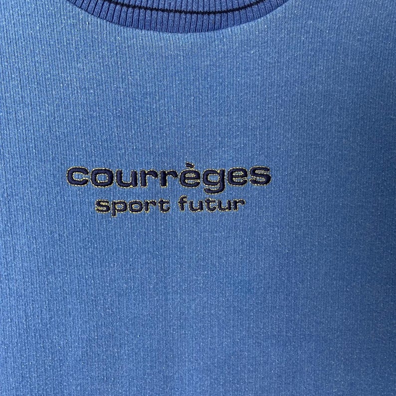 Puede incluir: Primer plano de una sudadera azul con el texto bordado "COURR&Egrave;GES sport futur" en hilo azul m&aacute;s oscuro. La sudadera tiene un cuello acanalado.