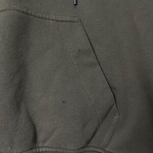 Puede incluir: Primer plano de una sudadera con capucha verde oscuro con un bolsillo de canguro. La tela tiene un aspecto ligeramente desgastado con peque&ntilde;as imperfecciones. El cord&oacute;n de la capucha es visible en la parte superior. La prenda est&aacute; hecha de un material suave y c&oacute;modo.
