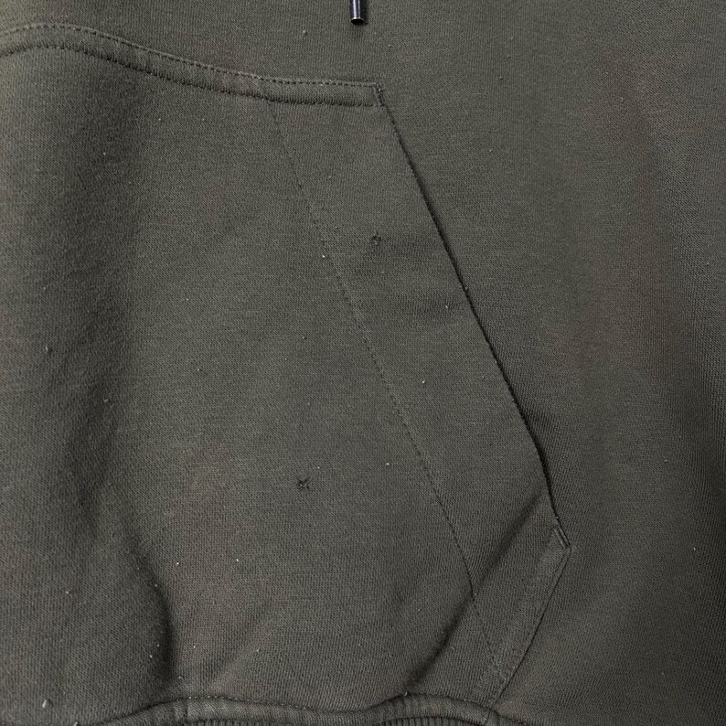 Puede incluir: Primer plano de una sudadera con capucha verde oscuro con un bolsillo de canguro. La tela tiene un aspecto ligeramente desgastado con peque&ntilde;as imperfecciones. El cord&oacute;n de la capucha es visible en la parte superior. La prenda est&aacute; hecha de un material suave y c&oacute;modo.