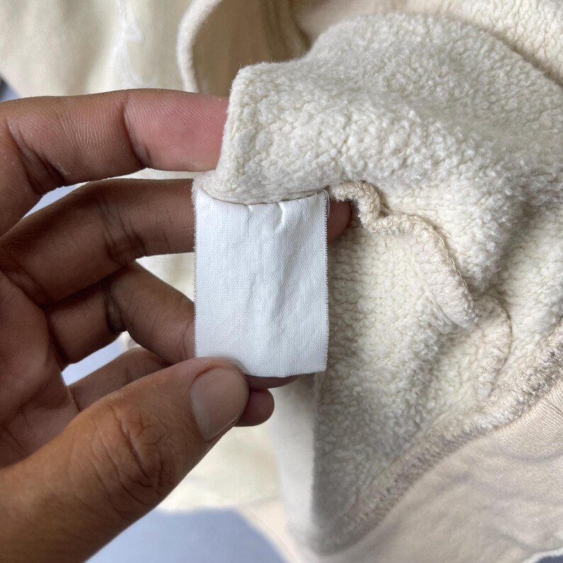 Puede incluir: Una sudadera de color crema con una textura interior suave y afelpada. Una etiqueta de cuidado rectangular blanca est&aacute; unida a la costura interior, mostrando una letra L tenue y parcialmente visible en el centro de la etiqueta.