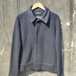 May include: A black textured jacket with a collar and button closure, hanging on a wooden hanger. The jacket has long sleeves and a slightly textured appearance. The jacket is displayed against a neutral background.