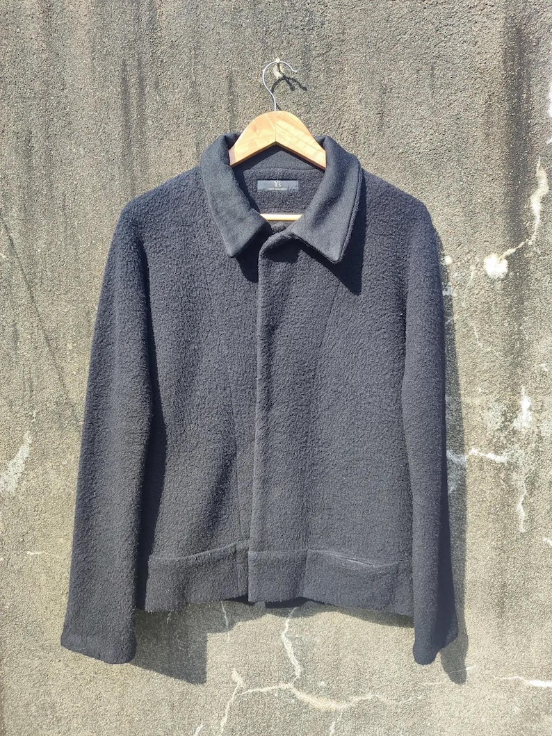 May include: A black textured jacket with a collar and button closure, hanging on a wooden hanger. The jacket has long sleeves and a slightly textured appearance. The jacket is displayed against a neutral background.