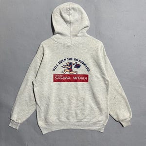 Puede incluir: Sudadera con capucha gris claro con un gráfico en la espalda. El gráfico presenta una figura corriendo con el texto "WILL HELP YOU GO FORWARD" y "SAGAWA MITAKA" en rojo. La sudadera tiene capucha con cordón y mangas largas.