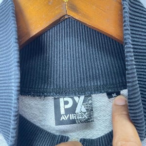 以下が含まれることがあります： 黒いリブ編みの襟が付いたスウェットシャツ。白い文字で「PX AVIREX」と書かれた正方形の黒いラベルと、「M」と書かれた小さな黒いサイズタグが付いています。襟の下にはグレーの質感のある裏地が見え、フロントにはジッパーが付いています。