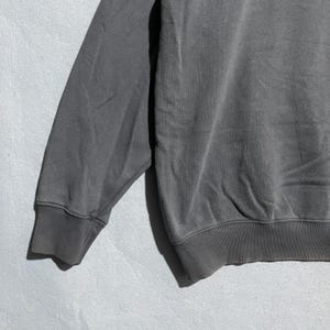 Puede incluir: Primer plano de una manga de sudadera gris. La manga est&aacute; hecha de una tela suave y texturizada y tiene un pu&ntilde;o acanalado. La sudadera es de color gris liso.