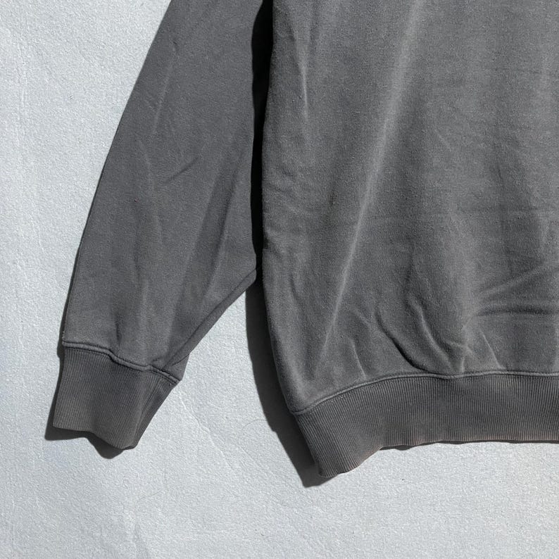 Puede incluir: Primer plano de una manga de sudadera gris. La manga est&aacute; hecha de una tela suave y texturizada y tiene un pu&ntilde;o acanalado. La sudadera es de color gris liso.