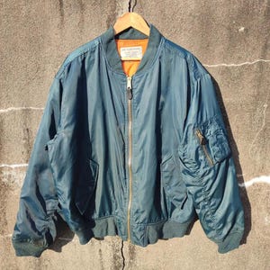 Puede incluir: Una chaqueta bomber azul oscuro con cierre de cremallera y un pequeño bolsillo en la manga izquierda. La chaqueta tiene una etiqueta que dice "ALPHA INDUSTRIES INC."