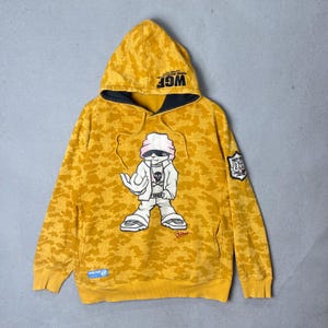 May include: A mustard yellow camouflage print hoodie featuring a graphic of a cartoon character wearing a pink beanie and white jacket. The hood interior is black with the text WGF printed on the edge. A shield patch with the number 03 is on the sleeve.