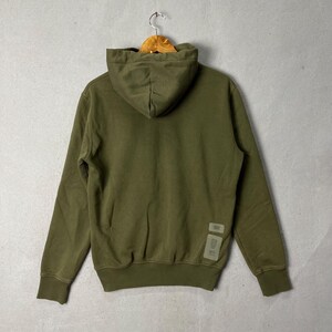 Puede incluir: Sudadera con capucha verde oliva con capucha con cord&oacute;n y mangas largas. La parte posterior de la sudadera presenta una peque&ntilde;a etiqueta cerca del dobladillo inferior. La sudadera con capucha est&aacute; colgada en una percha de madera contra una pared gris.