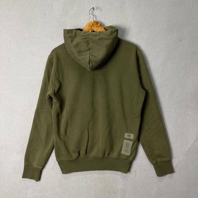 Puede incluir: Sudadera con capucha verde oliva con capucha con cord&oacute;n y mangas largas. La parte posterior de la sudadera presenta una peque&ntilde;a etiqueta cerca del dobladillo inferior. La sudadera con capucha est&aacute; colgada en una percha de madera contra una pared gris.