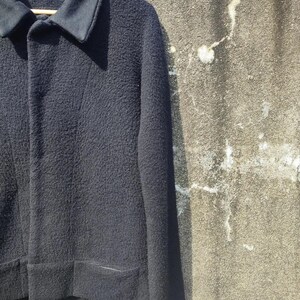 May include: A black jacket with a textured finish, a notched collar, and two front pockets. The jacket is suspended on a wooden hanger. The jacket is made of a thick material, possibly wool.