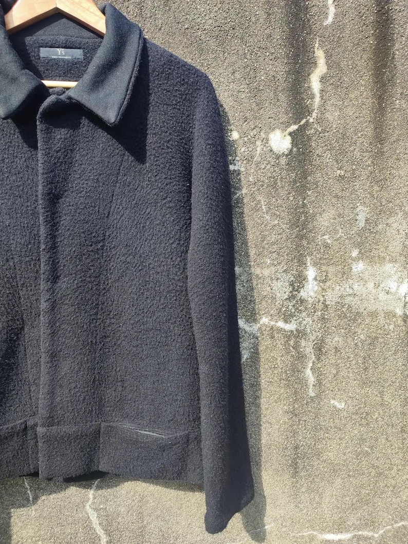 May include: A black jacket with a textured finish, a notched collar, and two front pockets. The jacket is suspended on a wooden hanger. The jacket is made of a thick material, possibly wool.