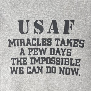 以下が含まれることがあります： グレーの霜降りTシャツで、黒い文字で「USAF MIRACLES TAKES A FEW DAYS THE IMPOSSIBLE WE CAN DO NOW.」とプリントされています。生地には目に見えるニットの質感があり、文字は胸元に太いブロック体のフォントで印刷されています。