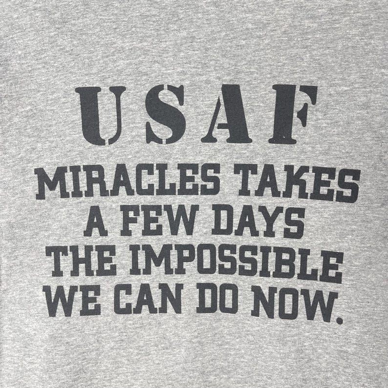以下が含まれることがあります： グレーの霜降りTシャツで、黒い文字で「USAF MIRACLES TAKES A FEW DAYS THE IMPOSSIBLE WE CAN DO NOW.」とプリントされています。生地には目に見えるニットの質感があり、文字は胸元に太いブロック体のフォントで印刷されています。