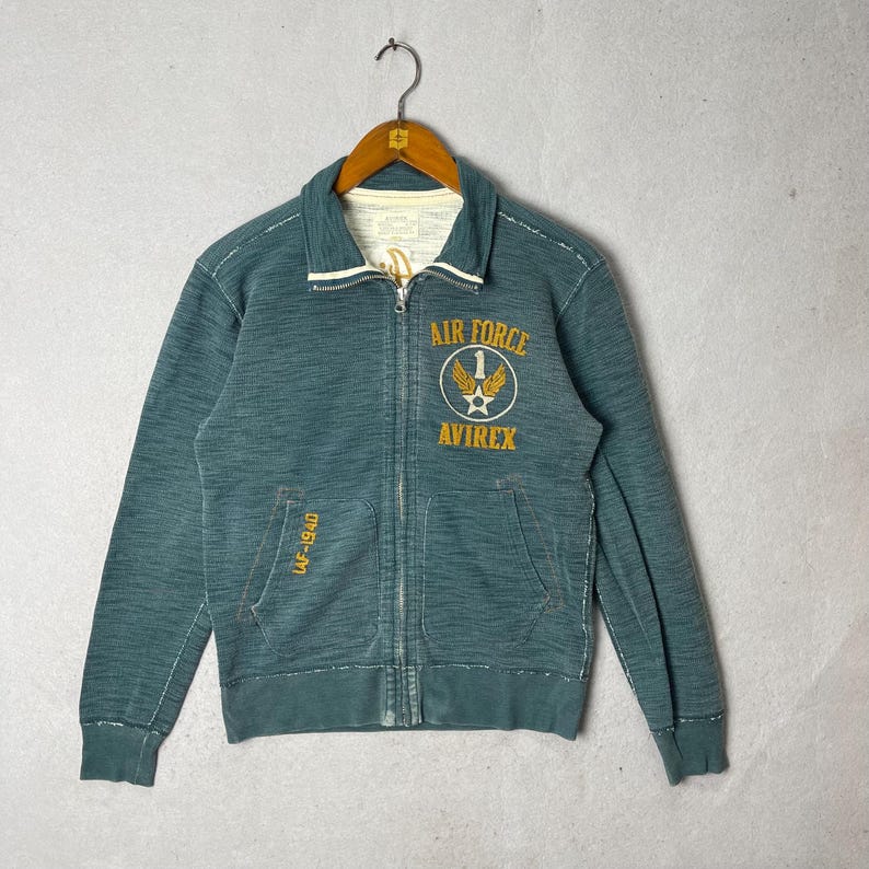 Peut inclure: Veste zipp&eacute;e bleu sarcelle avec l'inscription "AIR FORCE" et "AVIREX" en jaune. Col blanc et poche avec "LAF-1940". La veste est suspendue &agrave; un cintre m&eacute;tallique. V&ecirc;tement de style vintage.
