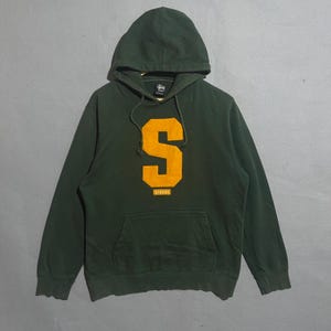 May include: Dark green hooded sweatshirt with a large yellow "S" graphic on the front. The word "Stussy" is printed in yellow below the letter. The sweatshirt has a front pocket and a drawstring hood.