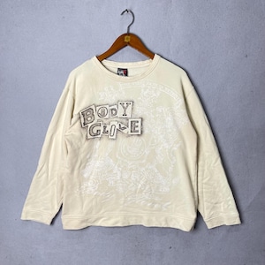 Puede incluir: Una sudadera de manga larga color crema con el texto "BODY GLIDE" en una fuente oscura y desgastada en la parte superior izquierda del pecho. La prenda incluye un gran dise&ntilde;o gr&aacute;fico blanco tenue en el frente y una peque&ntilde;a etiqueta de marca en el cuello.