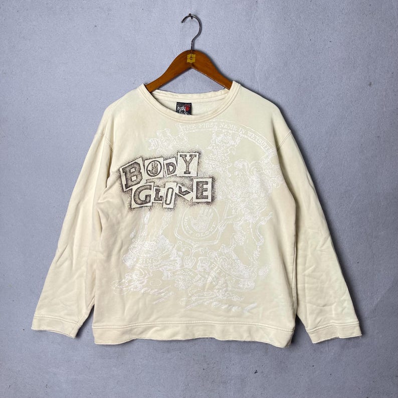 Puede incluir: Una sudadera de manga larga color crema con el texto "BODY GLIDE" en una fuente oscura y desgastada en la parte superior izquierda del pecho. La prenda incluye un gran dise&ntilde;o gr&aacute;fico blanco tenue en el frente y una peque&ntilde;a etiqueta de marca en el cuello.