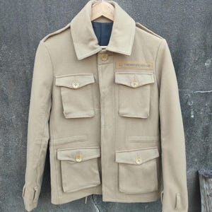 以下が含まれることがあります： ベージュ色のジャケット。前面に複数のポケットとボタン留めが付いています。ジャケットは幅広の襟とテキストの入った小さな長方形のパッチが付いています。木製のハンガーに掛けられています。