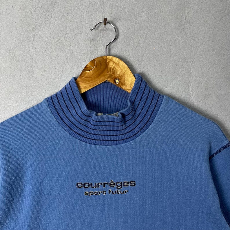 Puede incluir: Sudadera azul claro con cuello alto. El cuello tiene rayas horizontales negras. El nombre de la marca "courr&egrave;ges" y la frase "sport futur" est&aacute;n bordados en negro en la parte delantera. La sudadera est&aacute; colgada de una percha de madera.