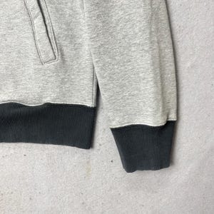 以下が含まれることがあります： ダークチャコールグレーのリブ編みの裾と、袖口に同色のリブを備えたライトグレーの長袖スウェットシャツ。前面左側に縦型のポケット口があり、滑らかなヘザー調の生地で構成されています。