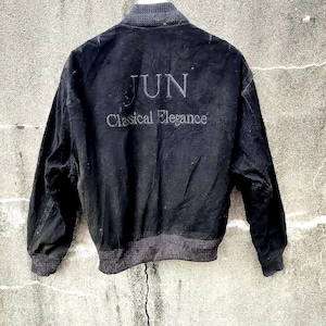 Pode incluir: Casaco bomber preto com gola e punhos canelados. A parte de trás do casaco apresenta o texto "JUN Classical Elegance" num design tonal. O casaco está pendurado contra uma parede de betão cinzento texturizado.