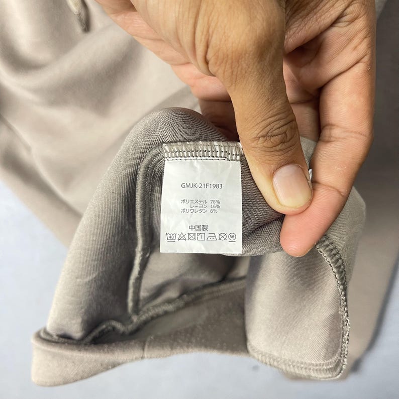 May include: Close-up of a taupe-colored garment tag held by a hand. The tag displays the text "GMJK-21F1983" and fabric composition details in Japanese. Washing instructions are also visible. The garment appears to be made of a soft, flexible material.