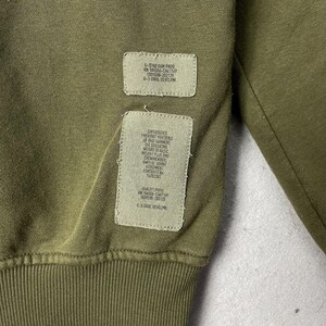 Puede incluir: Primer plano de una sudadera verde oliva con dos parches rectangulares cosidos. Los parches contienen texto blanco con detalles sobre la prenda, incluida la marca, el n&uacute;mero RN y otras especificaciones. La tela tiene una textura acanalada.