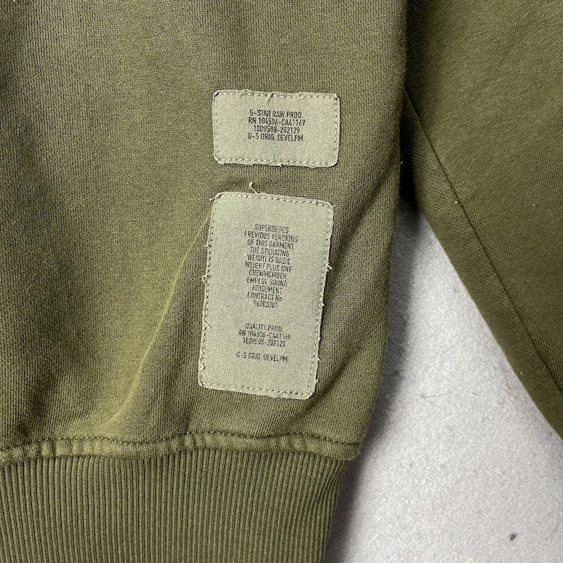 Puede incluir: Primer plano de una sudadera verde oliva con dos parches rectangulares cosidos. Los parches contienen texto blanco con detalles sobre la prenda, incluida la marca, el n&uacute;mero RN y otras especificaciones. La tela tiene una textura acanalada.