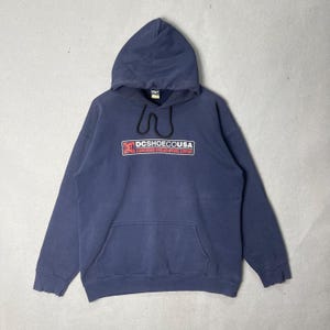 May include: A navy blue hooded sweatshirt with a front pocket and drawstring. The front features a rectangular logo with the text "DCSHOECOUSA" in white and red. The sweatshirt is made of a soft, worn material.