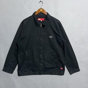 May include: Black Dickies zip-up jacket with a collar, small logo on the chest, and a red logo tag near the bottom. The jacket is hanging on a wooden hanger against a neutral background. The jacket has a full zipper and two front pockets.