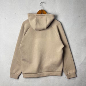 May include: A beige hoodie with a hood and long sleeves, hanging on a wooden hanger. The back of the hoodie is visible, showcasing its simple design and neutral color. The hoodie is made of a soft material.