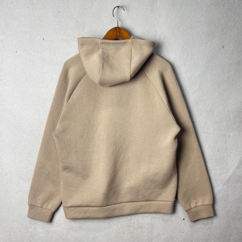 May include: A beige hoodie with a hood and long sleeves, hanging on a wooden hanger. The back of the hoodie is visible, showcasing its simple design and neutral color. The hoodie is made of a soft material.