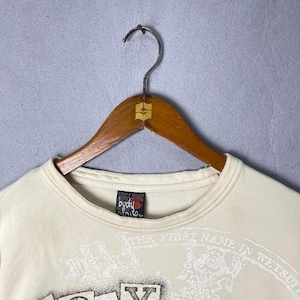 Puede incluir: Una sudadera de cuello redondo color crema con un estampado gr&aacute;fico en la parte delantera con el texto "THE FIRST NAME IN WETSUITS" y una etiqueta de la marca "Body Glove" en el interior del cuello, sobre una percha de madera con gancho met&aacute;lico.
