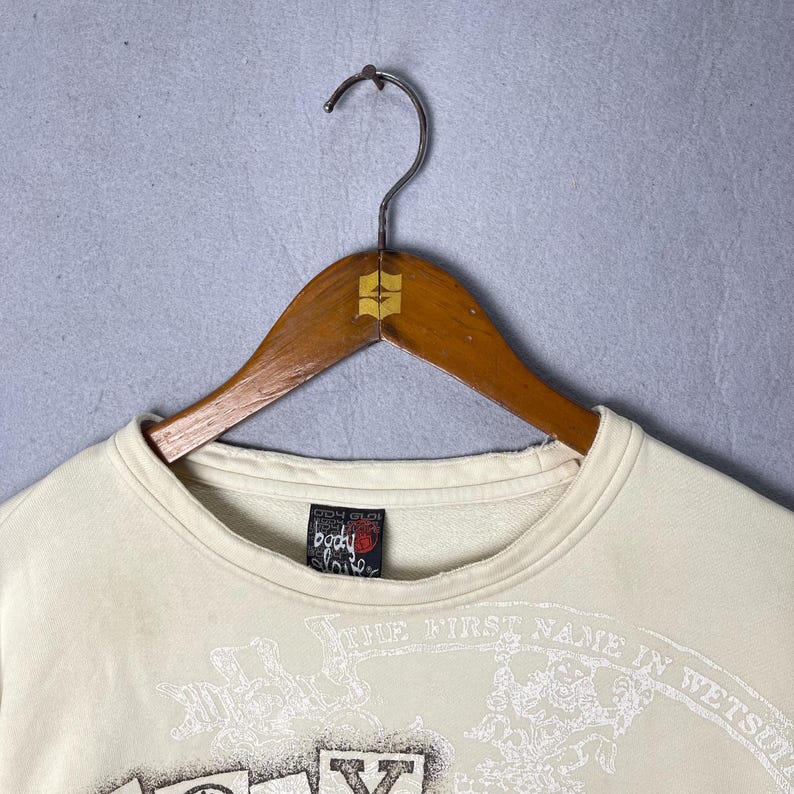 Puede incluir: Una sudadera de cuello redondo color crema con un estampado gr&aacute;fico en la parte delantera con el texto "THE FIRST NAME IN WETSUITS" y una etiqueta de la marca "Body Glove" en el interior del cuello, sobre una percha de madera con gancho met&aacute;lico.