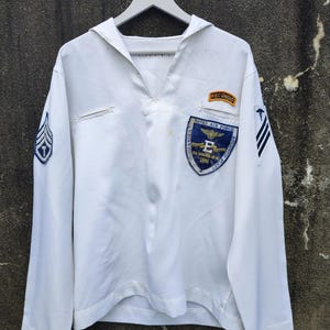 May include: White long-sleeved military shirt with a sailor collar. Features patches with text and insignia, including "RECONDO" and "NAVAL AIR FORCE". The shirt has blue and gold accents and a small pocket on the chest.