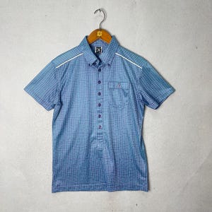 May include: Blue short-sleeved shirt with a grid pattern. The shirt has a button-down collar, a chest pocket, and white trim along the shoulders. It is hanging on a wooden hanger.