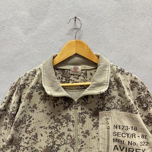 Puede incluir: Chaqueta con cremallera y estampado de camuflaje en tonos beige y marr&oacute;n. La chaqueta tiene cuello y pu&ntilde;os de canal&eacute;, cremallera frontal y bolsillo en el pecho con texto impreso. Cuelga de una percha de madera.