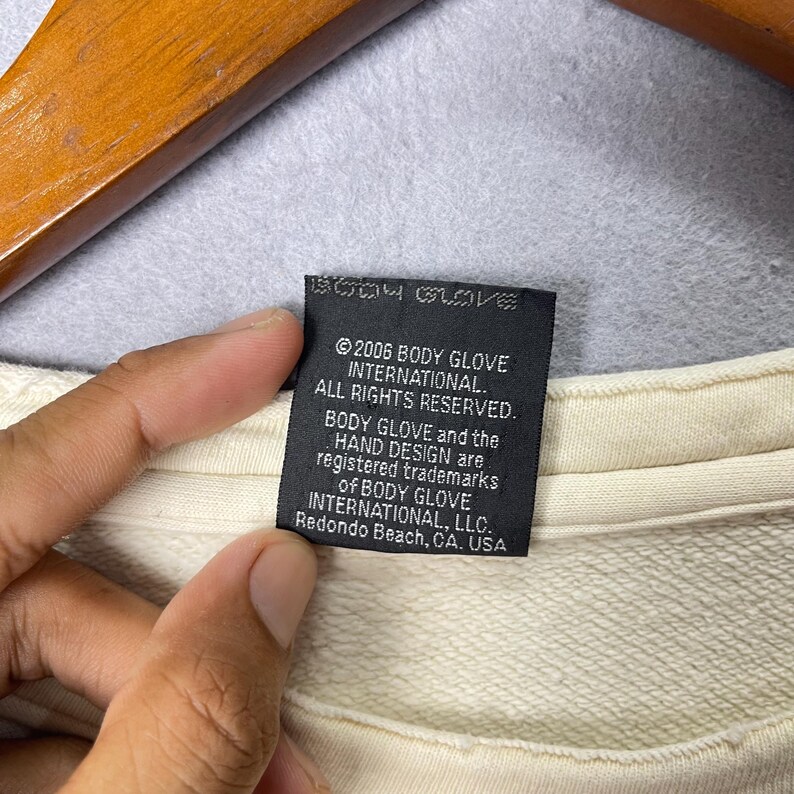Puede incluir: Una prenda de color crema con una etiqueta de tela rectangular negra con texto en blanco que dice: 2006 BODY GLOVE INTERNATIONAL. ALL RIGHTS RESERVED. BODY GLOVE and the HAND DESIGN are registered trademarks of BODY GLOVE INTERNATIONAL, LLC.