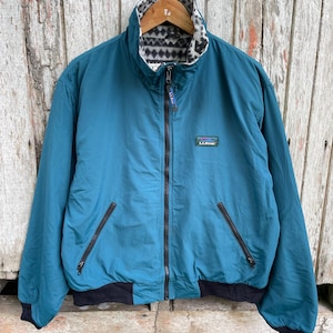 May include: Teal reversible jacket with a black and white patterned interior lining. The jacket has a full zipper, two front pockets, and a black ribbed hem and cuffs. The L.L.Bean logo is visible on the chest.