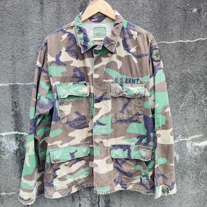 Könnte beinhalten: Tarnfarbene Militärjacke mit dem Text "U.S. ARMY" über den Taschen. Die Jacke hat ein grünes, braunes und violettes Tarnmuster. Ein Sternaufnäher ist am oberen Ärmel zu sehen. Die Jacke hängt an einem Holzkleiderbügel.