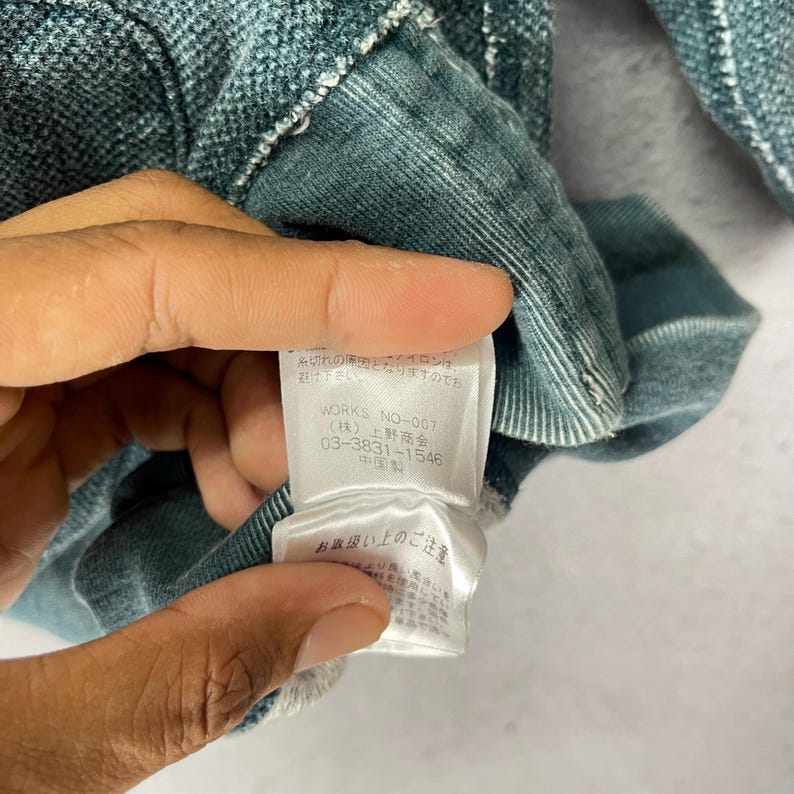 Peut inclure: Gros plan sur un v&ecirc;tement de couleur sarcelle avec un tissage textur&eacute;. Une &eacute;tiquette blanche est visible, avec du texte japonais et le nom de la marque "WORKS NO-007". L'&eacute;tiquette comprend &eacute;galement un num&eacute;ro de t&eacute;l&eacute;phone. Le tissu du v&ecirc;tement semble doux et l&eacute;g&egrave;rement us&eacute;.