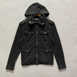 Könnte beinhalten: Schwarze Kapuzenjacke mit durchgehendem Reißverschluss, vier Vordertaschen und Schulterklappen. Die Jacke hat ein klassisches Design mit silberfarbenen Knöpfen und einem weichen, matten Finish. Die Kapuze ist befestigt und die Jacke ist aus glattem Stoff.
