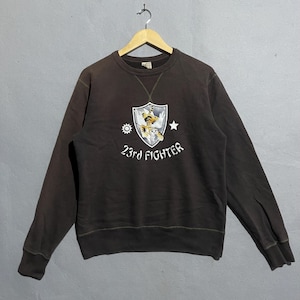 Può includere: Una felpa girocollo marrone con una grafica sul davanti. La grafica presenta uno scudo con una tigre, una stella e un disegno del sole, con il testo "23rd FIGHTER" sotto. La felpa è appesa a una gruccia di legno.