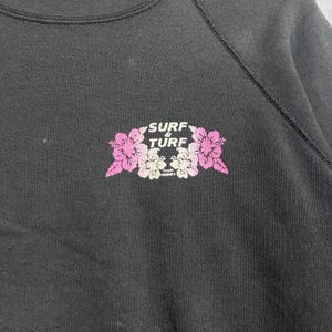 Op de afbeelding: Zwart sweatshirt met een bloemenprint met de tekst "Surf & Turf" in wit en roze. De afbeelding bevat roze en witte bloemen. De tekst "from Hawaii" staat eronder.