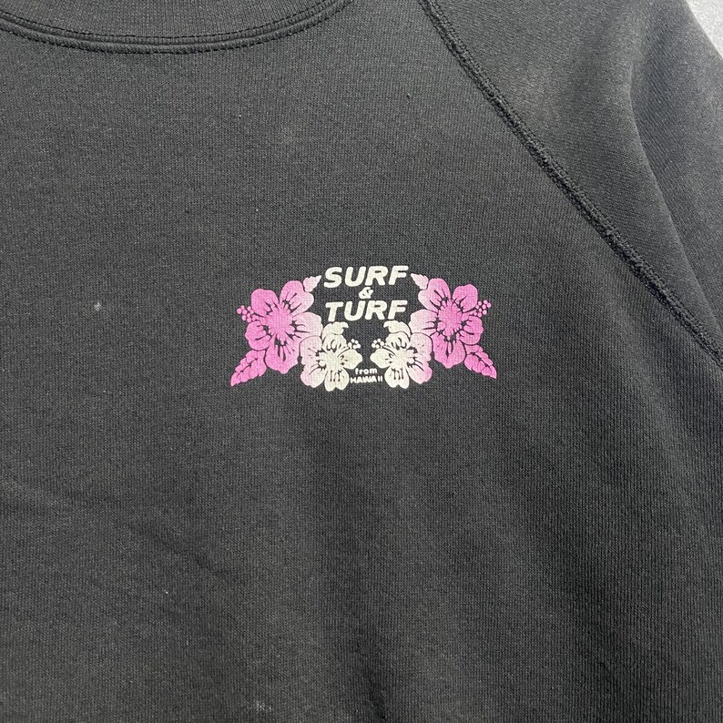 Op de afbeelding: Zwart sweatshirt met een bloemenprint met de tekst "Surf & Turf" in wit en roze. De afbeelding bevat roze en witte bloemen. De tekst "from Hawaii" staat eronder.