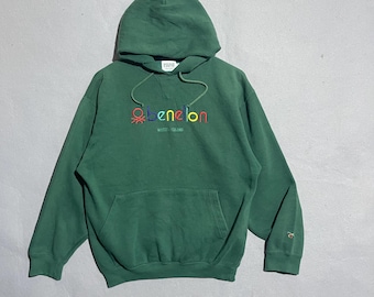 May include: Green hooded sweatshirt with a front pocket and drawstring. The word "benetton" is embroidered across the chest in colourful thread. A small logo is embroidered on the sleeve. The sweatshirt is made of a soft material.